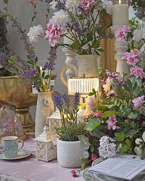 tea party table set up with artificial pastel flowers, table lamp, tea cup and saucer, jugs, lanterns, and candles