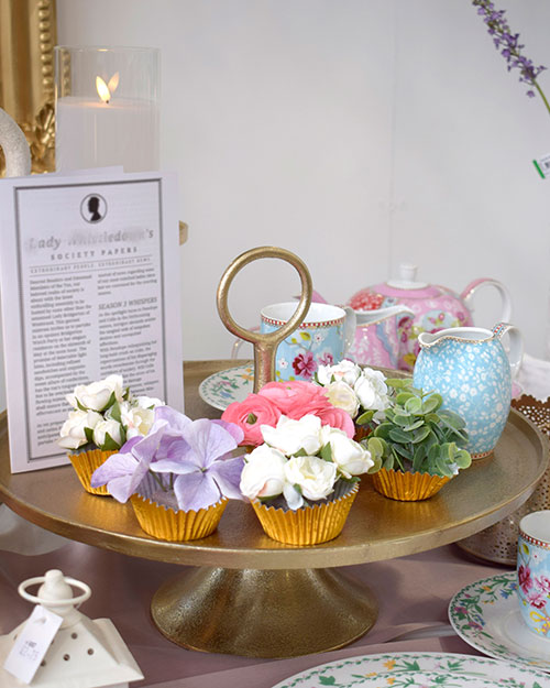 golden cupcake cups with faux flowers inside, tea cup and saucer, and jug on golden cake stand